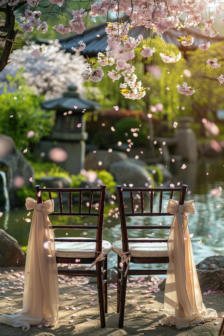 two chairs with sashes are set up in front of a pond and cherry blossom trees