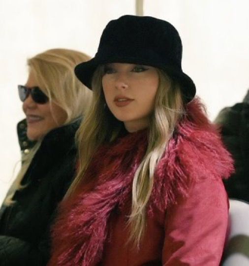 two women sitting next to each other in front of a white wall wearing black hats and fur vests