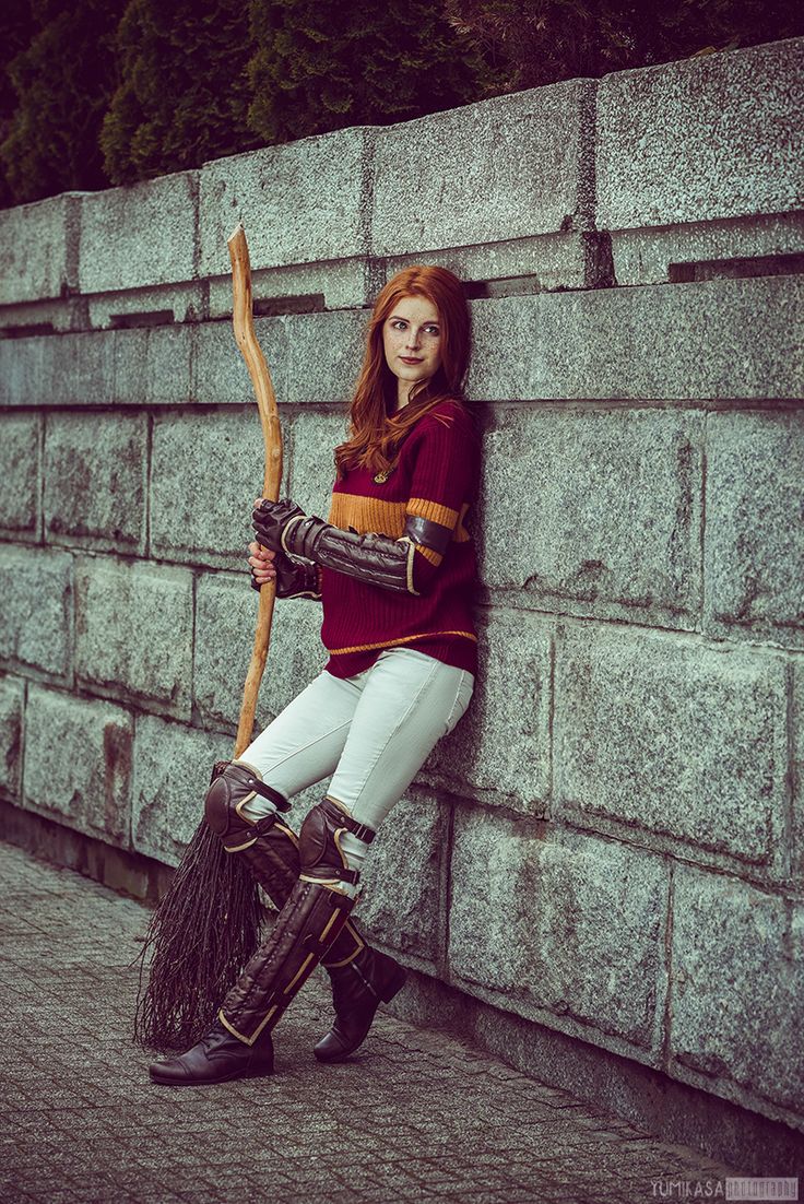 a woman dressed in medieval clothing leaning against a wall holding a broom and posing for the camera