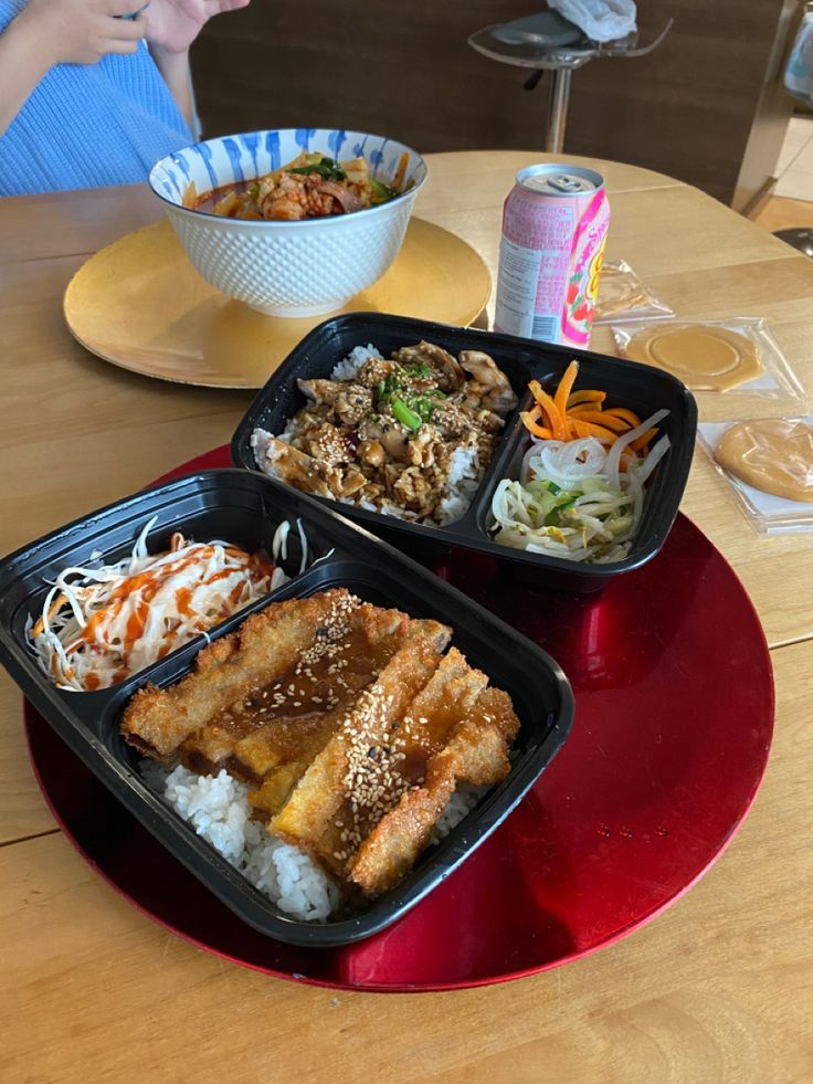 two trays of food sitting on top of a wooden table next to a bowl of rice