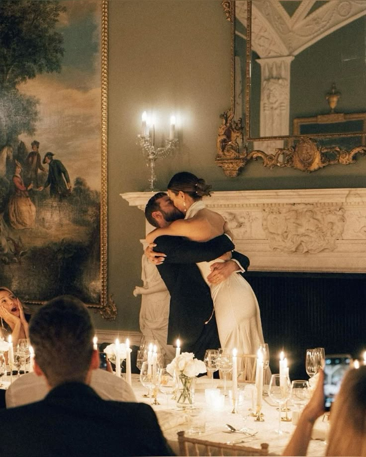 two people hugging each other at a dinner table with candles in front of the fireplace