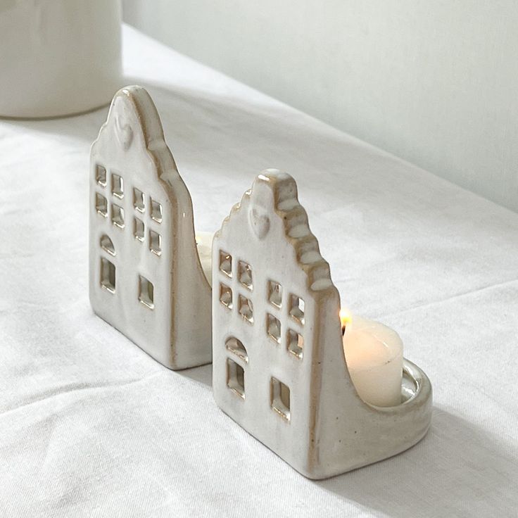 two white ceramic houses sitting on top of a bed next to a cup with a candle in it