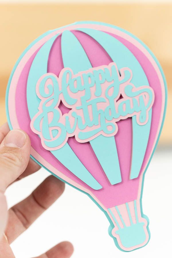 a hand holding up a pink and blue hot air balloon sticker that says happy birthday