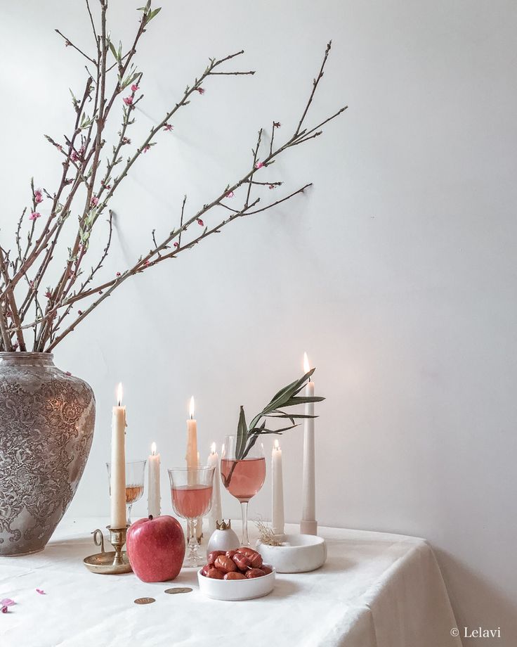 an arrangement of candles, fruit and flowers on a table with white walls in the background