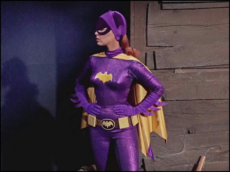 a woman dressed in purple and gold standing next to a wooden wall with her hands on her hips