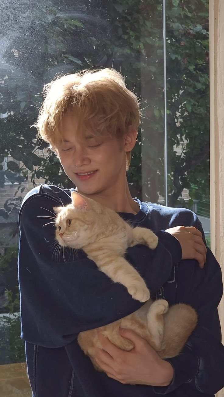 a boy holding a cat in his arms and smiling at the camera while standing next to a window