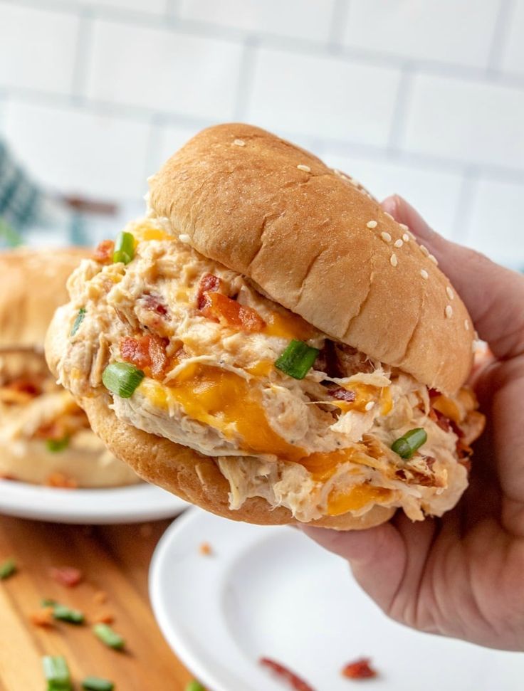 a hand holding up a chicken sandwich on a bun with cheese and green onions in the background