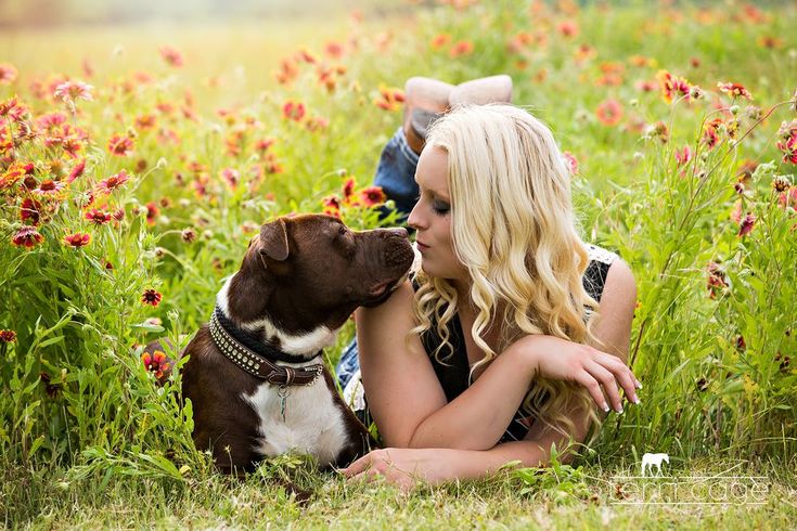 a woman is sitting in the grass with her dog and kissing her nose while she lays down