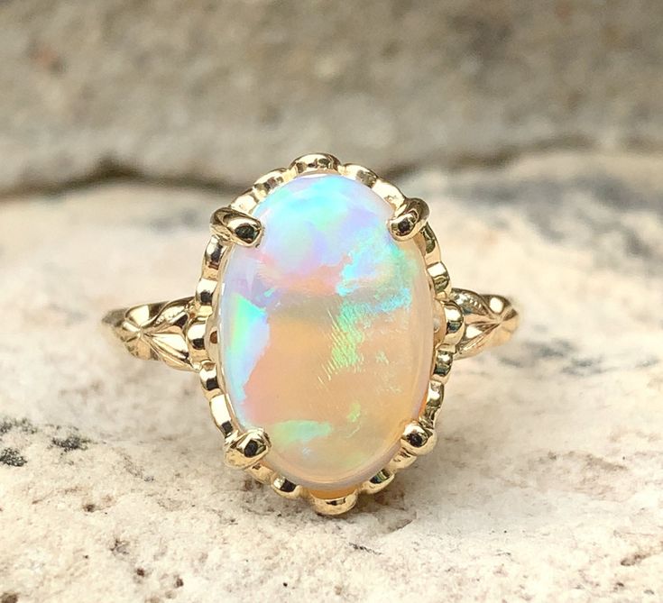 All of our jewelry is guaranteed authentic by our Graduate Gemologist of the Gemological Institute of America (GIA). 2.96ct Genuine Natural Opal 14k Yellow Gold Ring with Decorative Setting #J6080 14k yellow gold ring featuring an oval opal weighing 2.96 carats and measuring about 13mm x 9mm. The opal has blue-green play of color on a jelly/crystal semi-transparent base. The ring fits a size 7 finger and weighs 2.07 dwt. The mounting is vintage 1940s with a new head and stone. Breathtaking! SKU# Formal Iridescent Oval Ring, Elegant Oval Untreated Opal Ring, Elegant Untreated Oval Opal Ring, Elegant Iridescent Opal Ring For Anniversary, Elegant Iridescent Cabochon Opal Ring, Elegant Iridescent Opal Ring With Cabochon, Elegant Anniversary Opal Ring, Gia Certified Oval Cabochon Jewelry Gift, Elegant Oval Iridescent Opal Ring