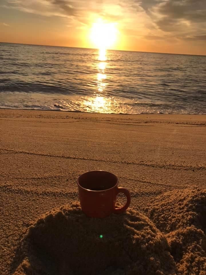 a coffee cup sitting on top of a sandy beach next to the ocean at sunset