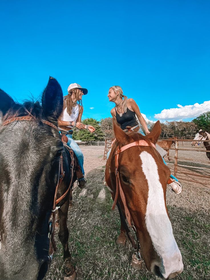 two women riding on the backs of brown and white horses in a corral area