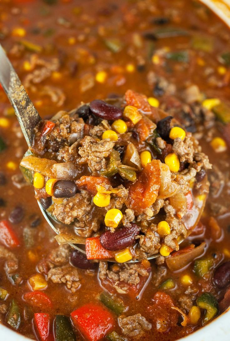 a spoon full of chili and ground beef soup with corn on the side in a white bowl