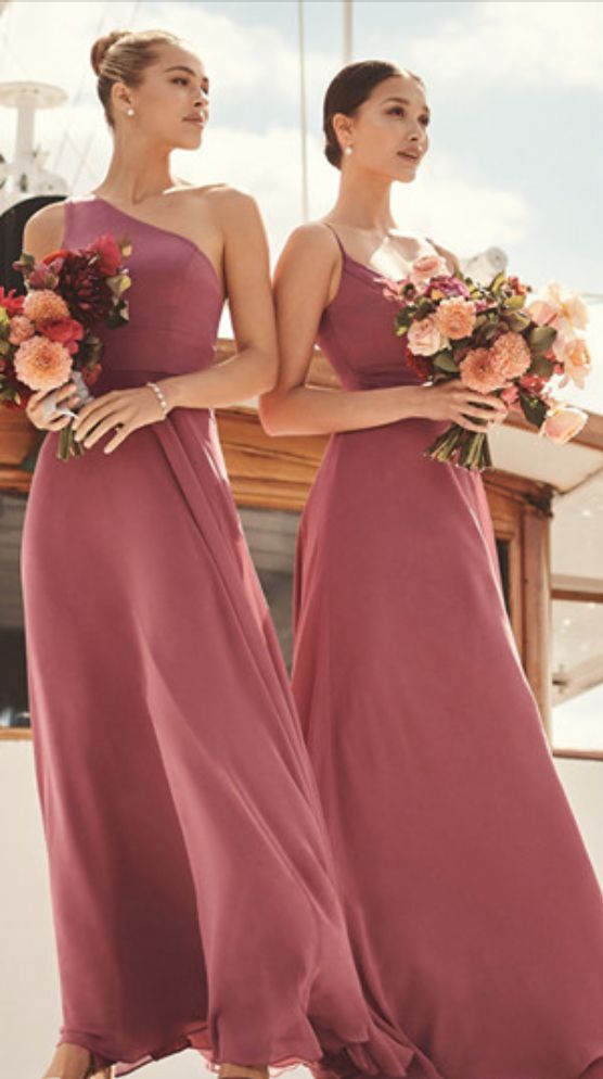 two women in long dresses standing next to each other with flowers on their heads and holding bouquets