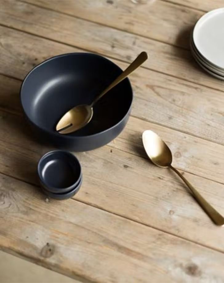 a wooden table topped with black bowls and gold spoons next to white plates on top of it