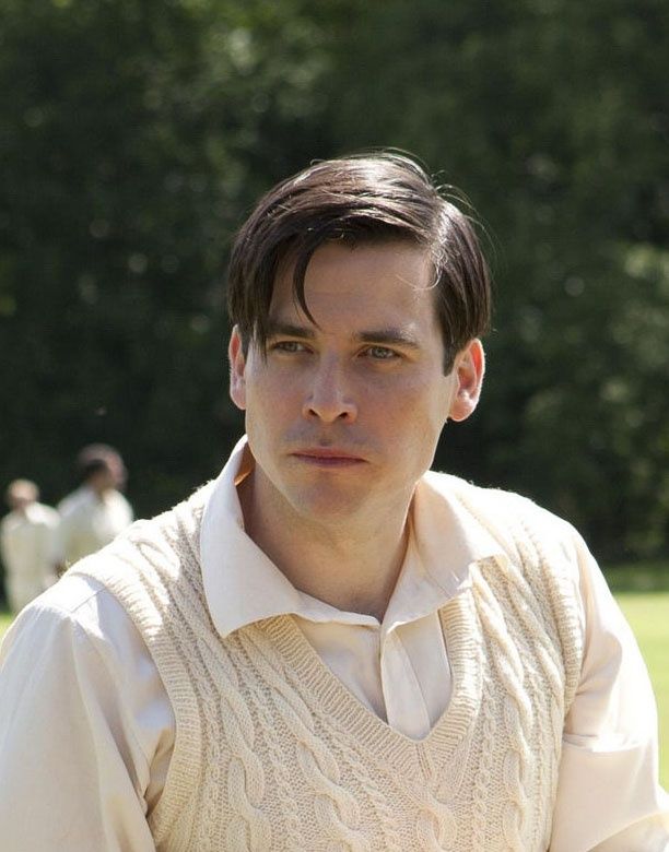 a man wearing a sweater and tie standing in front of some people on the grass