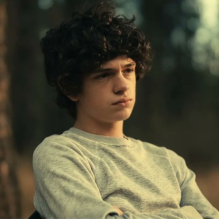 a young man with curly hair sitting in a chair looking off to the side while wearing a sweater