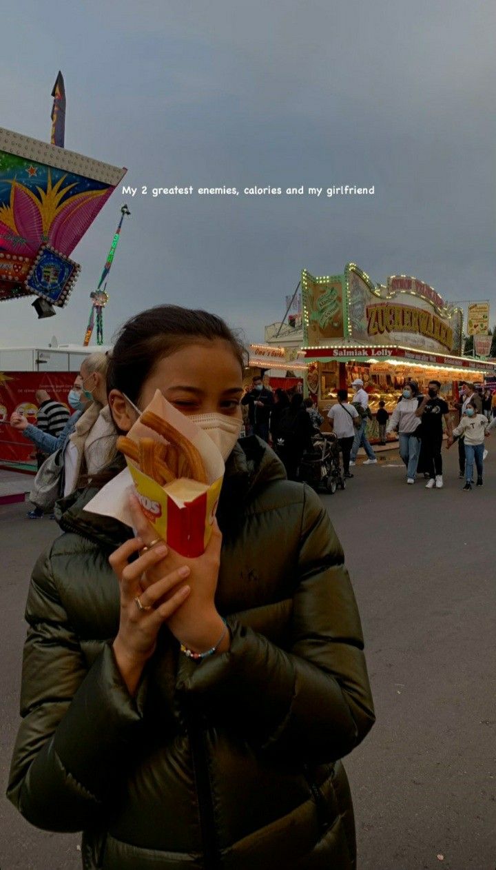 a woman in a puffy coat eating food at an amusement park with people walking around