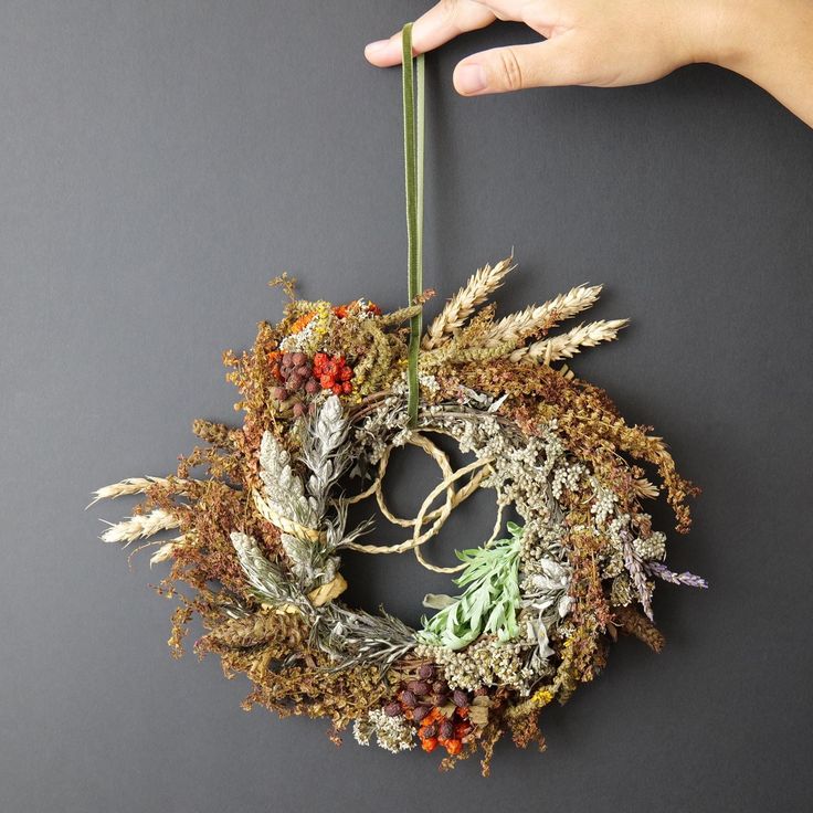 a hand holding a dried wreath on top of a gray surface with leaves and berries