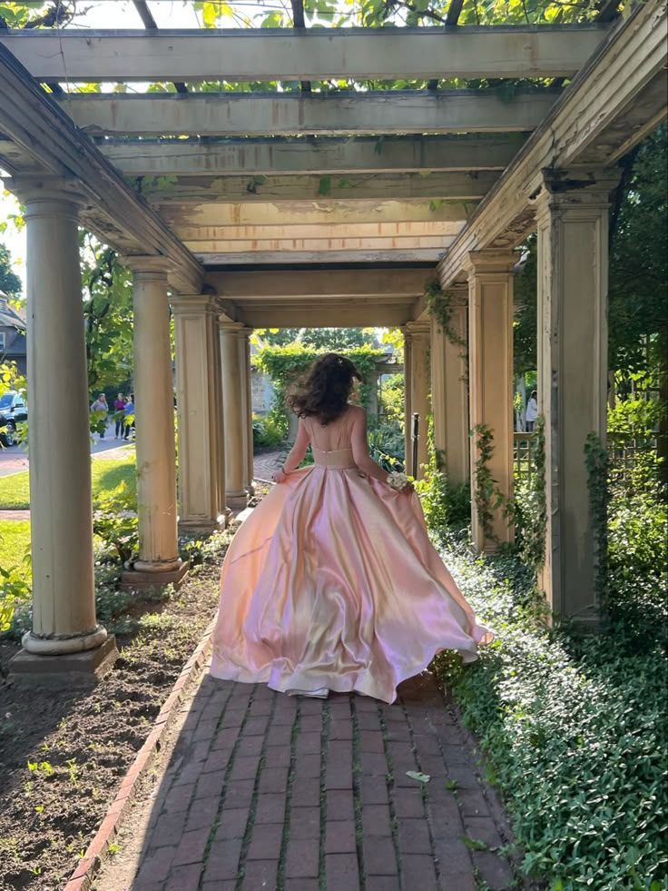 a woman in a long pink dress walking down a brick walkway under an arbored roof