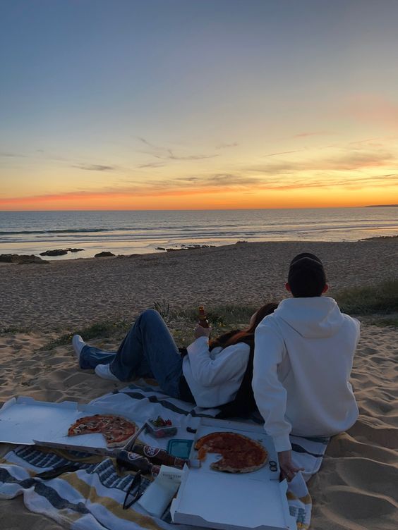 a man sitting on top of a beach next to the ocean eating pizza and watching the sun set