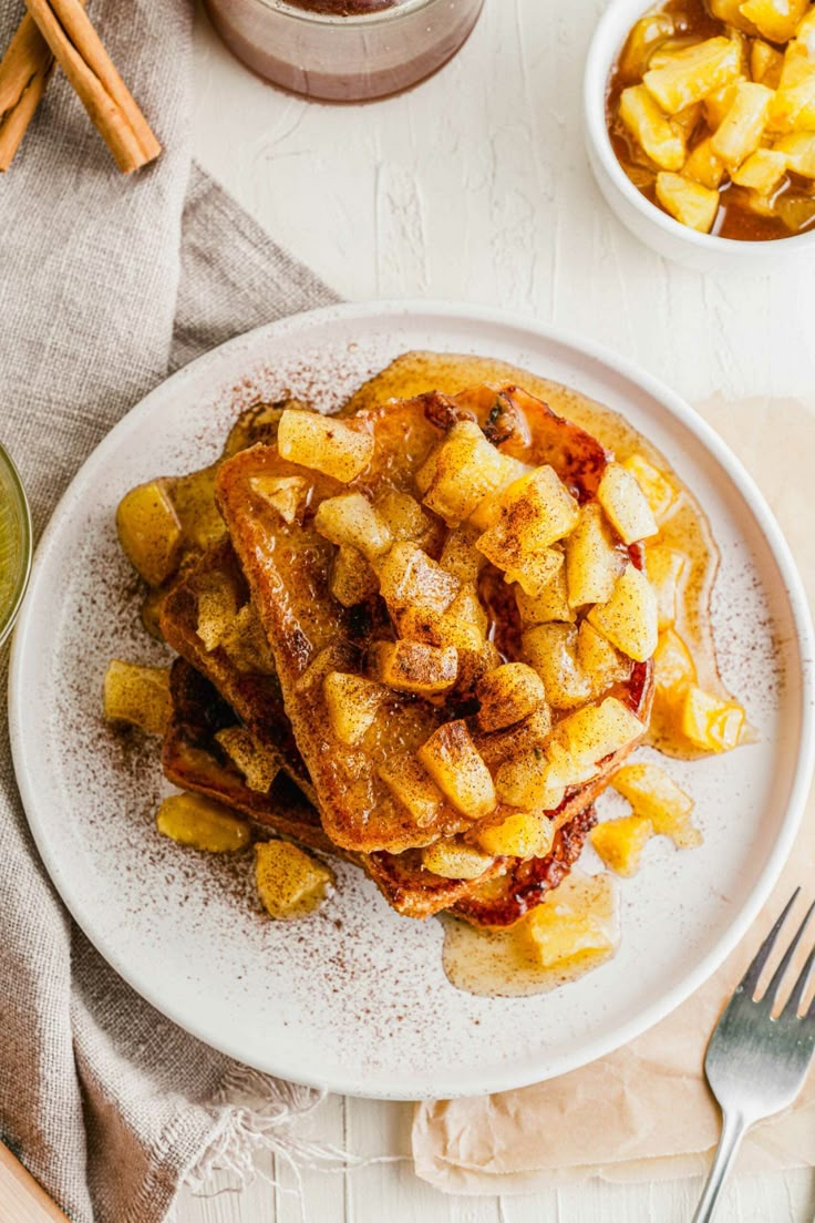 a white plate topped with french toast covered in syrup next to bowls of apples and cinnamon sticks