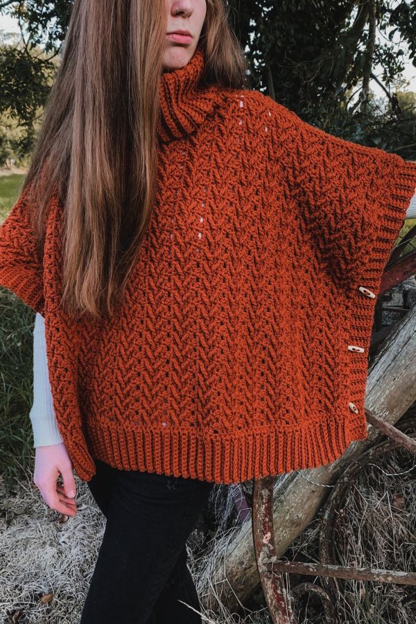 a woman wearing an orange knitted ponchy standing in front of a tree