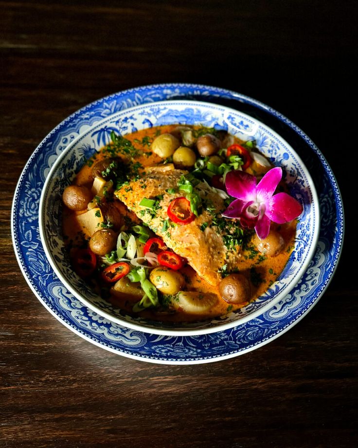 a blue and white bowl filled with food sitting on top of a wooden table next to a purple flower