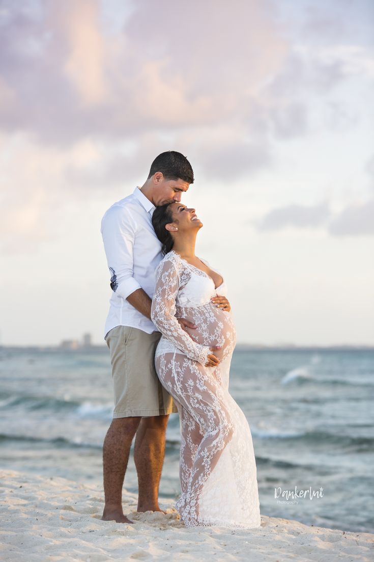 a man and woman standing on top of a sandy beach next to the ocean at sunset