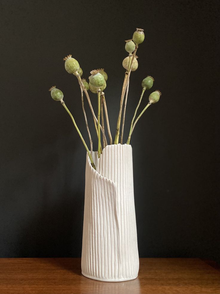 a white vase filled with green flowers on top of a wooden table next to a black wall