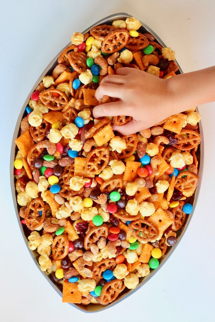 a tray filled with cereal and pretzels next to a hand reaching for it