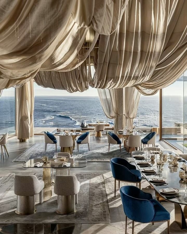 an ocean view restaurant with blue chairs and white drapes hanging from the ceiling, overlooking the water