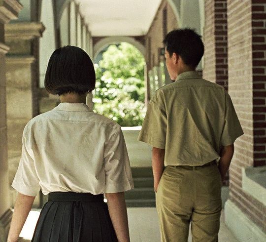 two people are walking down the hallway in an old fashioned school uniform, one is wearing glasses and the other wears a short sleeved shirt