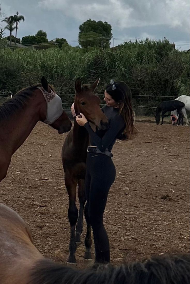 a woman standing next to two horses on a dirt field with trees in the background