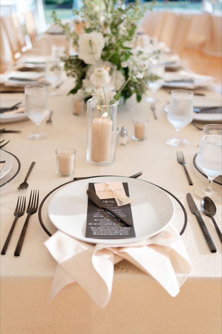 neutral wedding reception design showing a rectangular guest table with textured ivory and gold table linens under a black and clear rimmed charger, bone white dinner plate, black matte flatware and a black dinner menu with taupe place cards and a champagne tassel. Also showing taupe pillar and votive candles with a white & green floral centerpiece in the distance. Champagne Black And White Wedding, White And Champagne Wedding, Champagne And Black Wedding, Black And Cream Wedding, Black And Champagne Wedding Colors, Black White And Champagne Wedding, Cream Wedding Theme, Black Wedding Table, Wedding Reception Chairs