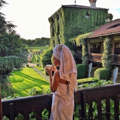 a woman standing on a balcony next to a lush green field and building with ivy covered walls