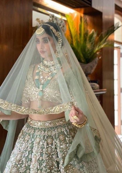 a woman in a green lehenga and veil with flowers on the skirt is standing near a potted plant