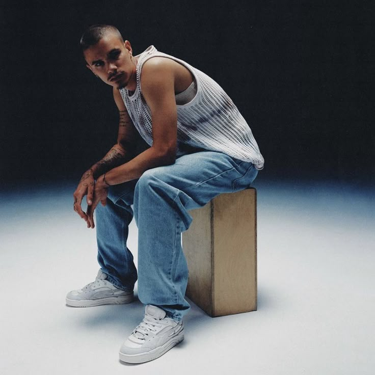 a man sitting on top of a wooden block with his hands in his pockets and wearing white sneakers