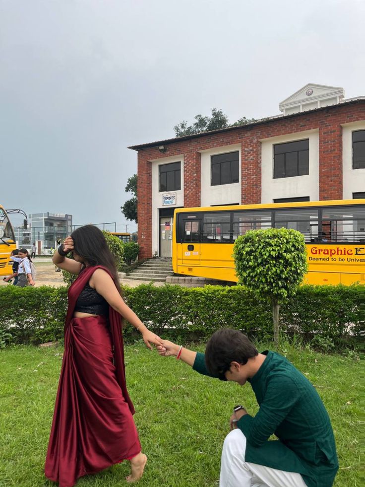 a man kneeling down next to a woman on top of a lush green field with a yellow bus in the background