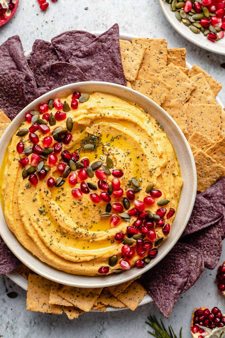 a white bowl filled with hummus and pomegranates surrounded by tortilla chips