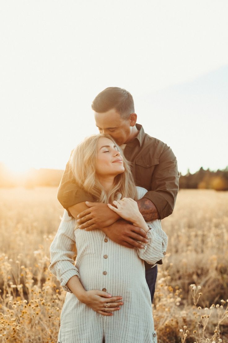 a pregnant woman hugging her husband in a field at sunset with the sun shining on her