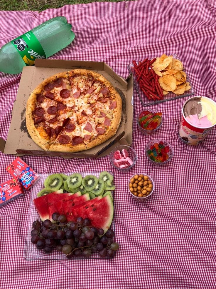 a picnic table with pizza, watermelon and other snacks on the blanket next to it