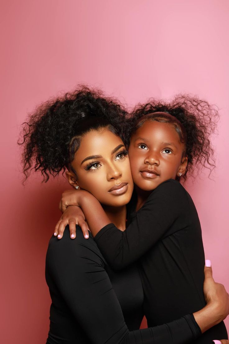 two young women hugging each other in front of a pink background, one is wearing black