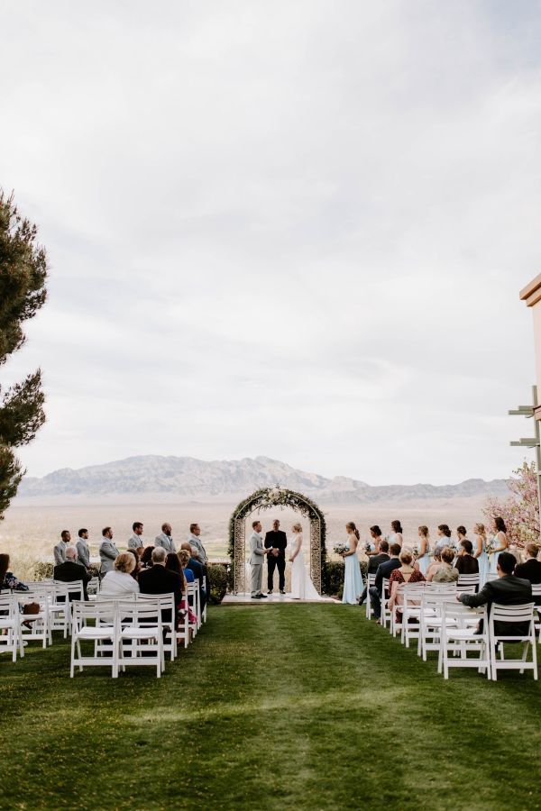 an outdoor ceremony with white chairs and people standing in front of the arch on the grass