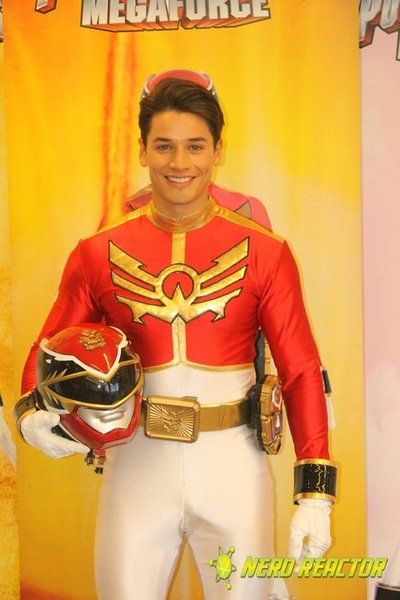 a man in a red and gold uniform holding a helmet on his right hand, standing next to a sign that says megaforce