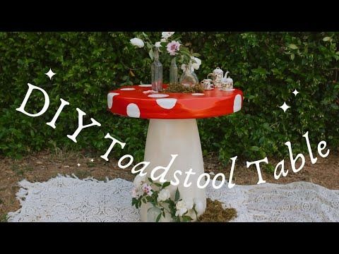 a red and white mushroom table with flowers on it in front of some shrubbery