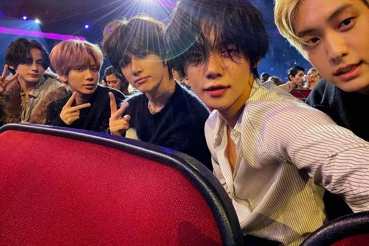 group of young men sitting in front of a red chair at a concert with one pointing to the side