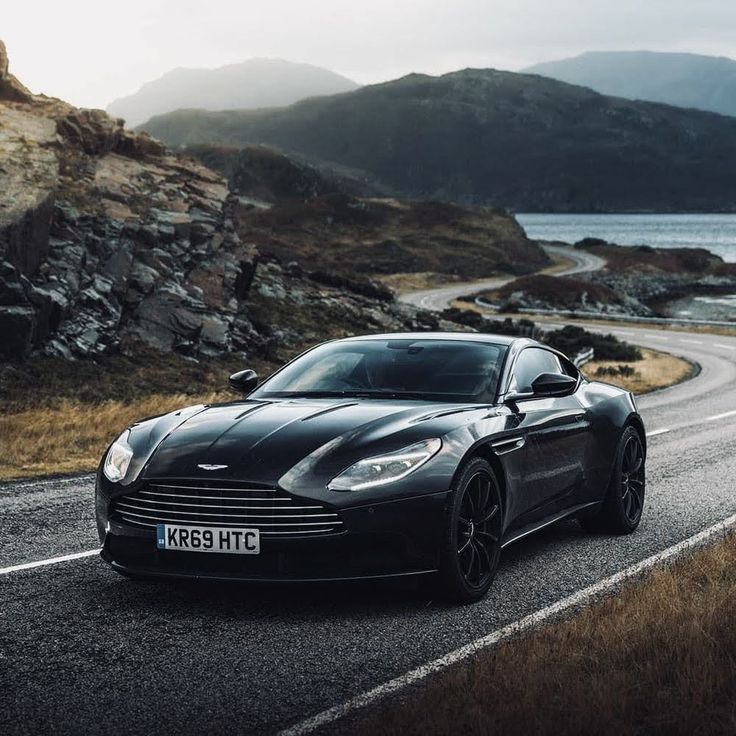 a black sports car driving down a road next to the ocean with mountains in the background