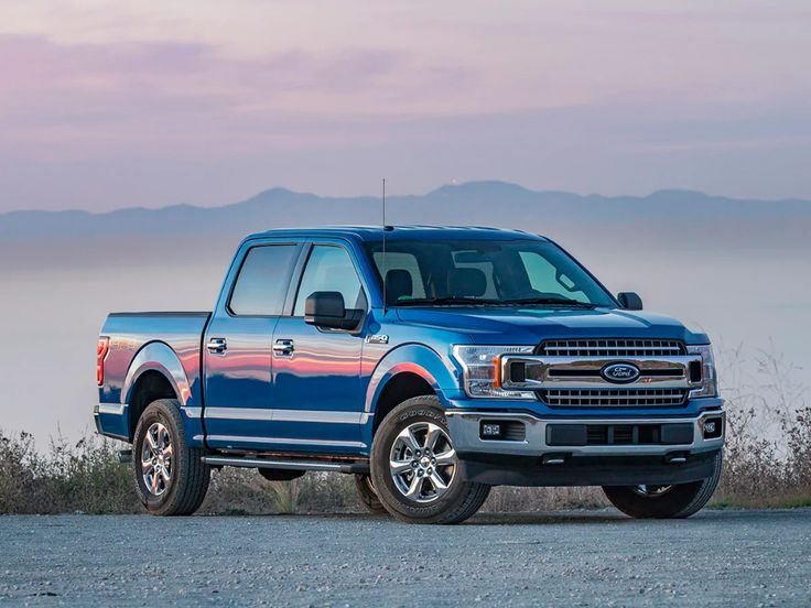 a blue truck parked on the side of a road near some grass and bushes with mountains in the background