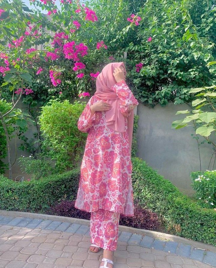 a woman wearing a pink hijab standing in front of some bushes and flowers