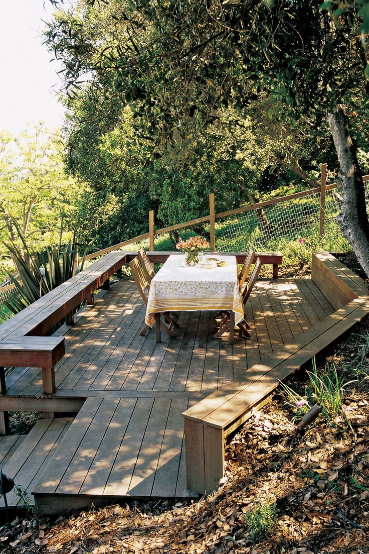 a table and benches on a wooden deck in the woods with trees around it,